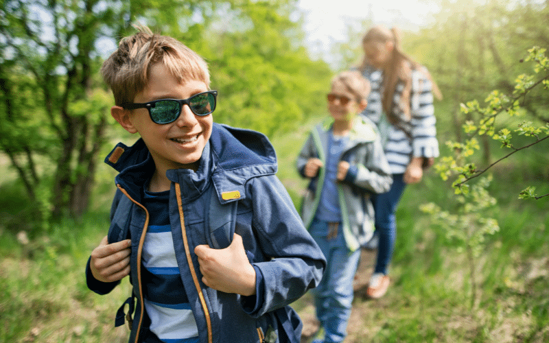 Children hiking with backpacks, walking sticks, and water bottles in a lush forest with a clear trail and colorful wildflowers